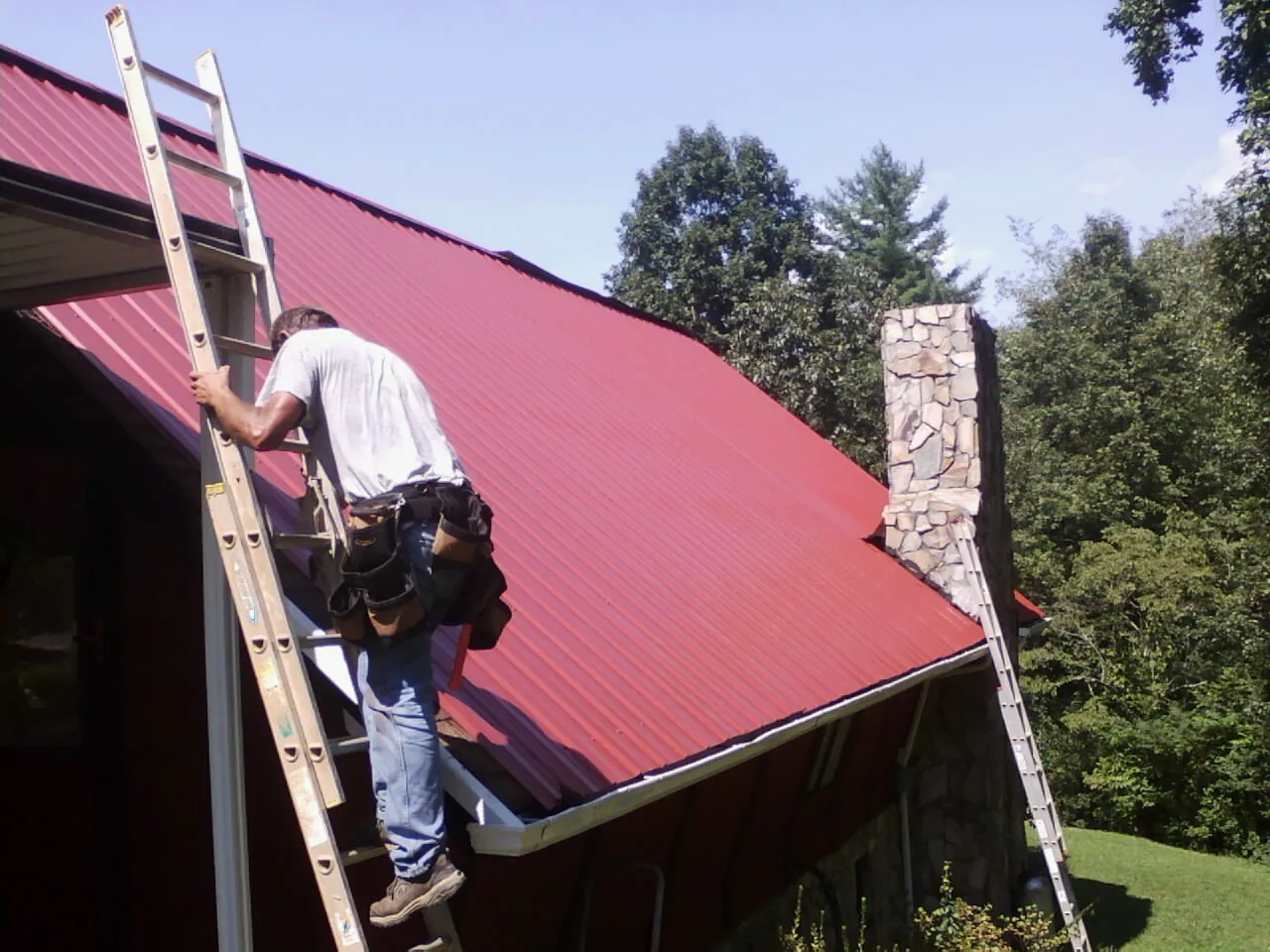 Roofer on a ladder beside a red metal roof and stone chimney — Soffit & Fascia Repair by Roofing Brothers in Bellvue